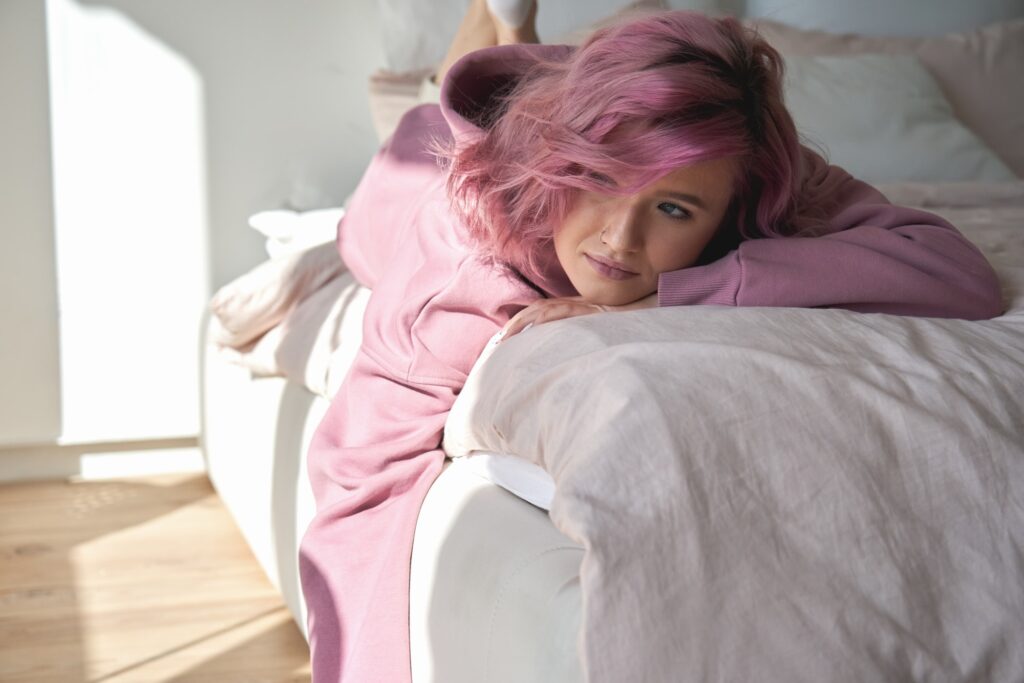 Depressed pensive teen girl introvert pink hair lying on bed looking away.