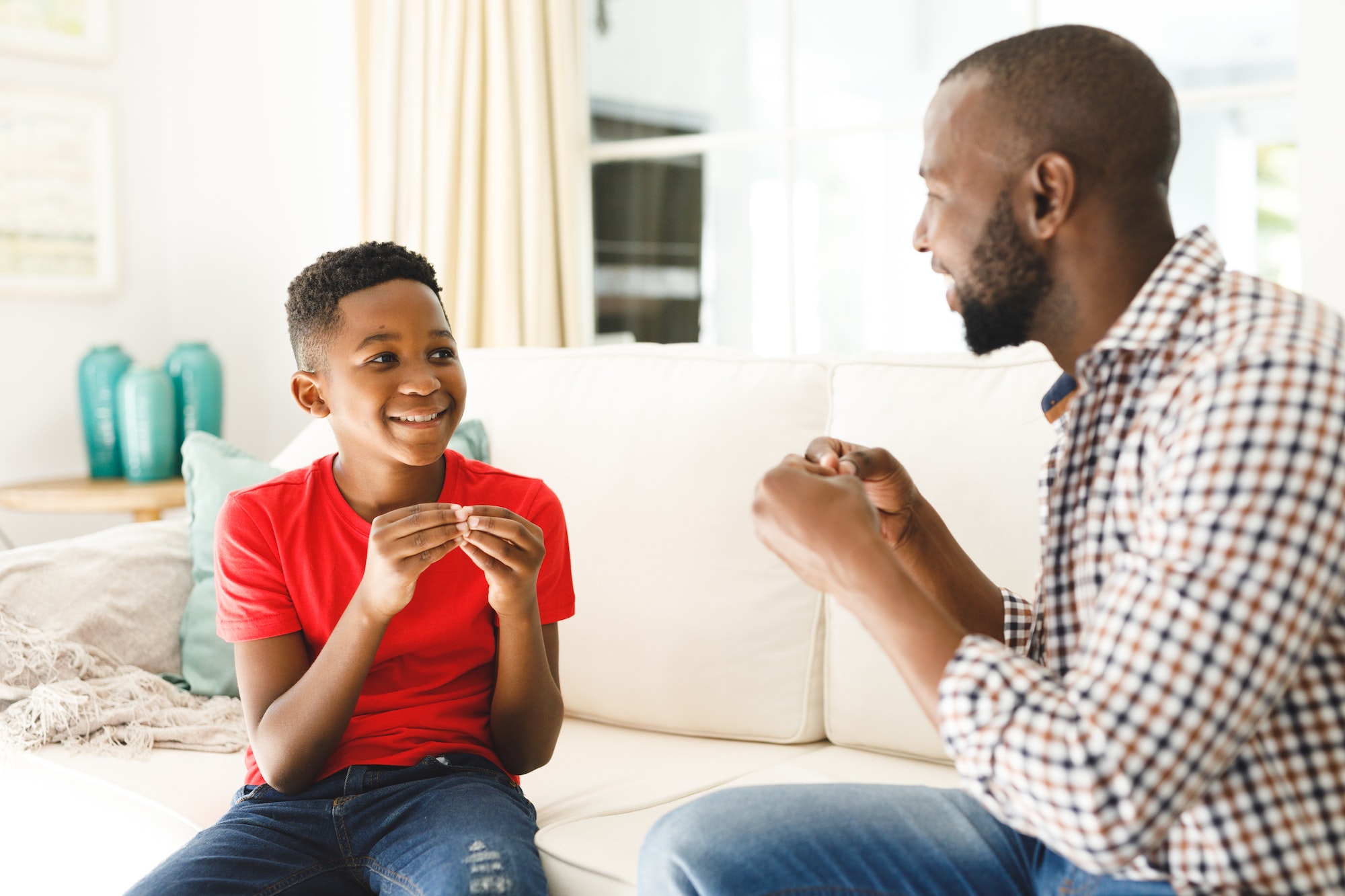 Happy african american father with son sitting on couch in living room talking sign language